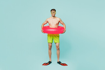 Full body confident young man wear green shorts swimsuit relax near hotel pool hold in hand inflatable rubber ring flippers isolated on plain blue background. Summer vacation sea rest sun tan concept.