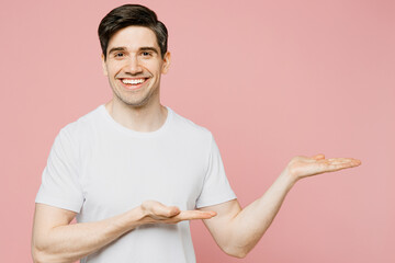 Wall Mural - Young smiling happy caucasian man wear white t-shirt casual clothes point hands arms aside on workspace area mock up isolated on plain pastel light pink background studio portrait. Lifestyle concept.