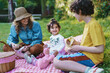 © Lomb - Joyful Mother Enjoying Picnic with Kids at Park - Caucasian mother with a straw hat, enjoying a sunny day having a picnic with her two cheerful children. They're interacting and eating bananas.