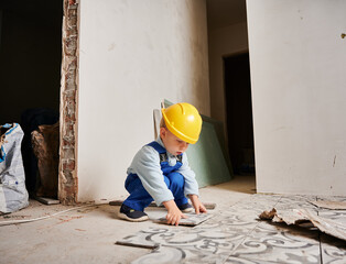 Little boy construction worker installing ceramic tile on the floor in apartment under renovation. Kid wearing safety helmet and work overalls while playing at home.