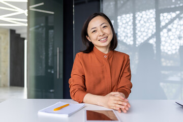 Portrait of young Asian successful business woman, female worker at workplace inside office smiling and looking at camera, video call online consultation listening to interlocutor with smile.