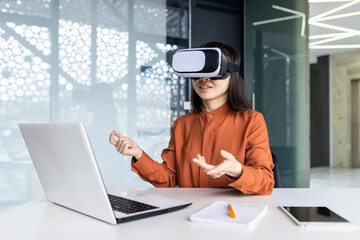 Asian woman wearing virtual reality glasses sitting at a desk in a workspace inside the office, businesswoman in online meeting talking remotely with colleagues, virtual meeting simulation.