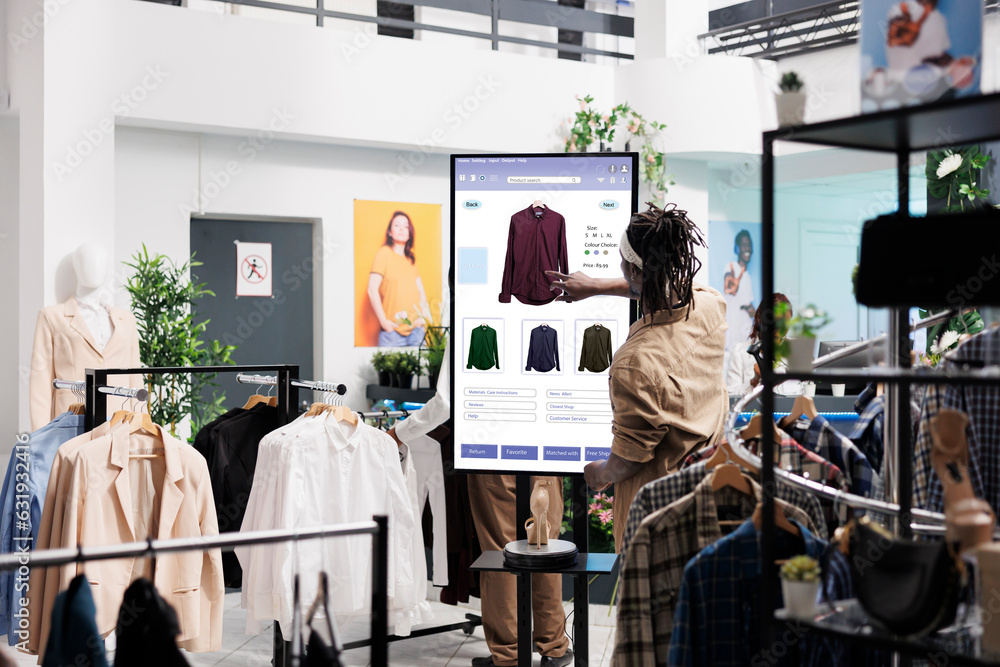Client using touch screen board in trendy clothing store, shopping for fashion collection items in shop. Young buyer buying clothes on interactive monitor, self ordering concept.
