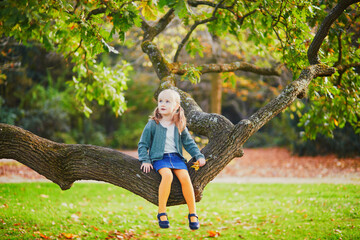 Wall Mural - Cute preschooler girl sitting on a tree branch in park or forest