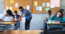 Male Teacher Helps Teen Male High School Student With Lesson In The Background. Classmates Focused On The Task At Hand.