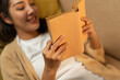 © Chanakon - Close up female hands holding book turning the page. Young woman sitting on cozey couch sofa reading book in living room with warm light at night. People female stay home part time relax at home.