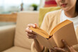 © Chanakon - Close up female hands holding book turning the page. Young woman sitting reading book on cozey couch sofa in living room with sun light morning. People female stay home part time relax at home.