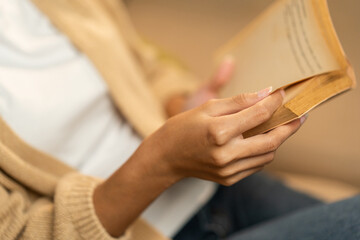 Close up female hands holding book turning the page. Young woman sitting on cozey couch sofa reading book in living room with warm light at night. People female stay home part time relax at home.