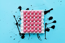Still Life Of White Carton With Bright Pink Eggs Standing On Blue Board With Abstract Black Paint Strokes, Directly Above View 