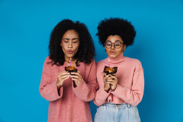 Wall Mural - Two happy african women blowing out candles on birthday cupcakes while standing isolated over blue wall