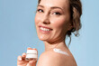 A smiling adult woman is taking care of her skin and holding a jar of cream and looking at the camera against a blue background