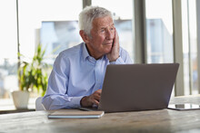 Portrait Of Pensive Senior Man Sitting At Table With Laptop Looking At Distance