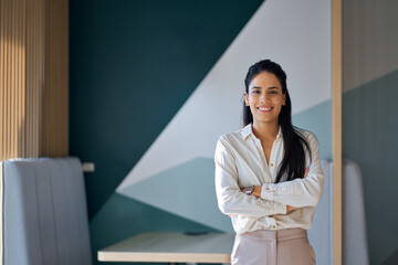 Wall Mural - Confident professional young latin business woman company employee, lady executive manager, female worker or entrepreneur looking at camera standing arms crossed in modern office, portrait.