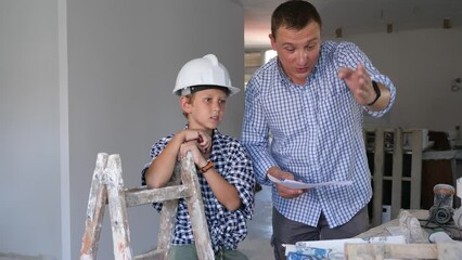 Wall Mural - Father and his young son wearing protective hardhats discussing work plan in construction site in apartment. 