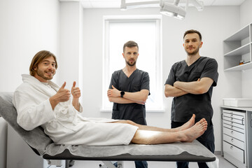 Wall Mural - Two confident surgeons talk with a young male patient lying on a couch with trauma in medical office. Concept of traumatology and rehabilitation