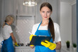 © Volha - Close-up of a beautiful young cleaning woman looking into the camera, checking a list on a paper tablet, watching the cleaning team work. Team of professional cleaners in uniform cleans the kitchen