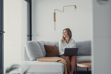 Portrait of one young attractive blonde woman using laptop pc computer on couch relaxing surfing the net at home looking at the window waiting package