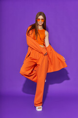 Wall Mural - Young fashion woman in orange pants orange top orange shirt on violet background. Platform slides sandals, orange sunglasses.