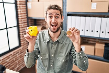 Wall Mural - Hispanic man with beard working at small business ecommerce holding piggy bank and bitcoin celebrating crazy and amazed for success with open eyes screaming excited.