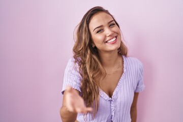 Wall Mural - Young hispanic woman standing over pink background smiling cheerful offering palm hand giving assistance and acceptance.