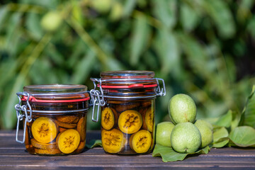 Wall Mural - Tincture of green walnuts in a glass jars on a wooden table in a summer garden. Sliced unripe walnuts in alcohol in a jar, to prepare homemade tincture, closeup