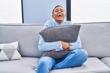 Sticker - African american woman sitting on the sofa at home hugging pillow smiling and laughing hard out loud because funny crazy joke.