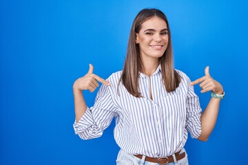 Canvas Print - Hispanic young woman standing over blue background looking confident with smile on face, pointing oneself with fingers proud and happy.