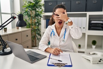 Canvas Print - Young hispanic woman wearing doctor uniform and stethoscope peeking in shock covering face and eyes with hand, looking through fingers with embarrassed expression.