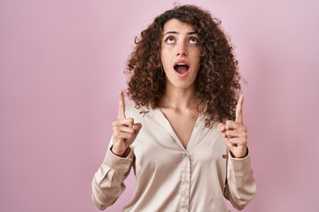 Sticker - Hispanic woman with curly hair standing over pink background amazed and surprised looking up and pointing with fingers and raised arms.