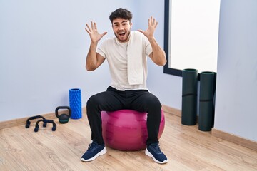 Wall Mural - Hispanic man with beard sitting on pilate balls at yoga room celebrating crazy and amazed for success with arms raised and open eyes screaming excited. winner concept