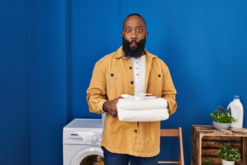 Poster - African american man holding clean towels at laundry room making fish face with mouth and squinting eyes, crazy and comical.