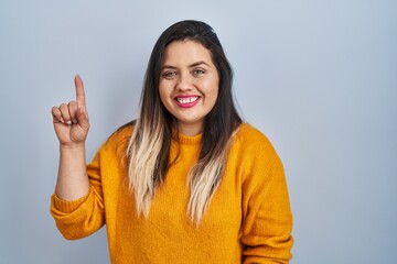 Wall Mural - Young hispanic woman standing over isolated background showing and pointing up with finger number one while smiling confident and happy.