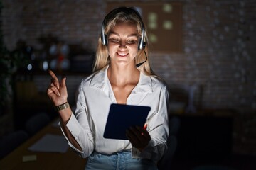 Poster - Young blonde woman working at the office at night with a big smile on face, pointing with hand finger to the side looking at the camera.