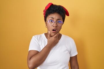 Wall Mural - Young african american woman standing over yellow background looking fascinated with disbelief, surprise and amazed expression with hands on chin