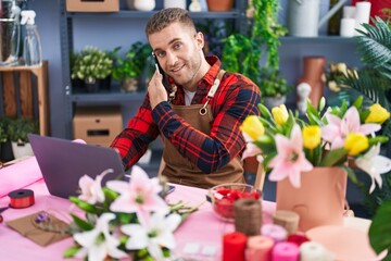 Wall Mural - Young caucasian man florist talking on smartphone using laptop at flower shop