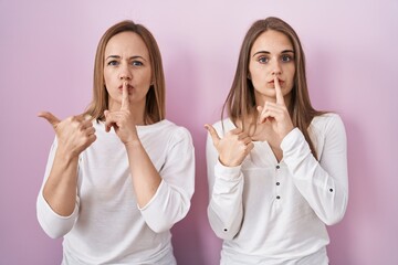 Canvas Print - Middle age mother and young daughter standing over pink background asking to be quiet with finger on lips pointing with hand to the side. silence and secret concept.