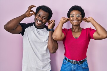 Poster - Young african american couple standing over pink background smiling pulling ears with fingers, funny gesture. audition problem