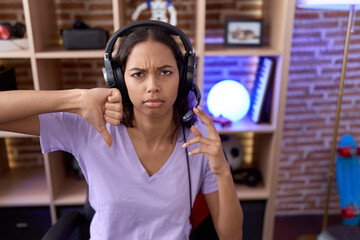 Sticker - Young hispanic woman playing video games wearing headphones with angry face, negative sign showing dislike with thumbs down, rejection concept