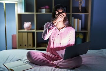 Sticker - Young hispanic woman using computer laptop on the bed covering eyes with hand, looking serious and sad. sightless, hiding and rejection concept