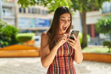 Wall Mural - Young beautiful hispanic woman using smartphone with surprise expression at park