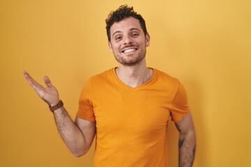 Sticker - Young hispanic man standing over yellow background smiling cheerful presenting and pointing with palm of hand looking at the camera.