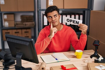 Sticker - Hispanic man working at small business ecommerce holding open banner serious face thinking about question with hand on chin, thoughtful about confusing idea