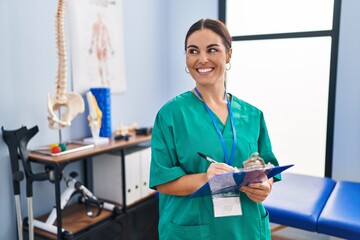 Canvas Print - Young beautiful hispanic woman pysiotherapist smiling confident writing on document at rehab clinic