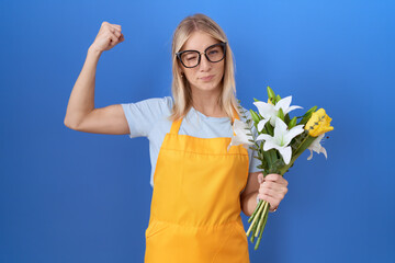 Poster - Young caucasian woman wearing florist apron holding flowers strong person showing arm muscle, confident and proud of power