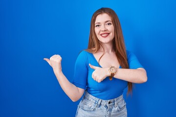 Poster - Redhead woman standing over blue background pointing to the back behind with hand and thumbs up, smiling confident