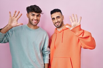 Young hispanic gay couple standing over pink background showing and pointing up with fingers number ten while smiling confident and happy.