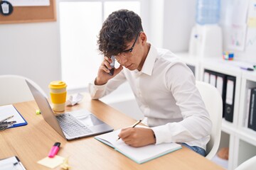 Poster - Young hispanic teenager business worker talking on smartphone writing on notebook at office