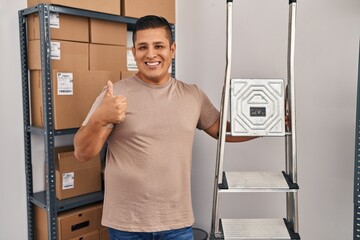 Wall Mural - Hispanic young man working at small business ecommerce holding ladders smiling happy and positive, thumb up doing excellent and approval sign
