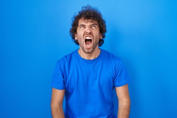 Sticker - Hispanic young man standing over blue background angry and mad screaming frustrated and furious, shouting with anger. rage and aggressive concept.