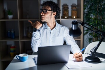 Poster - Hispanic man working at the office at night smiling with happy face looking and pointing to the side with thumb up.